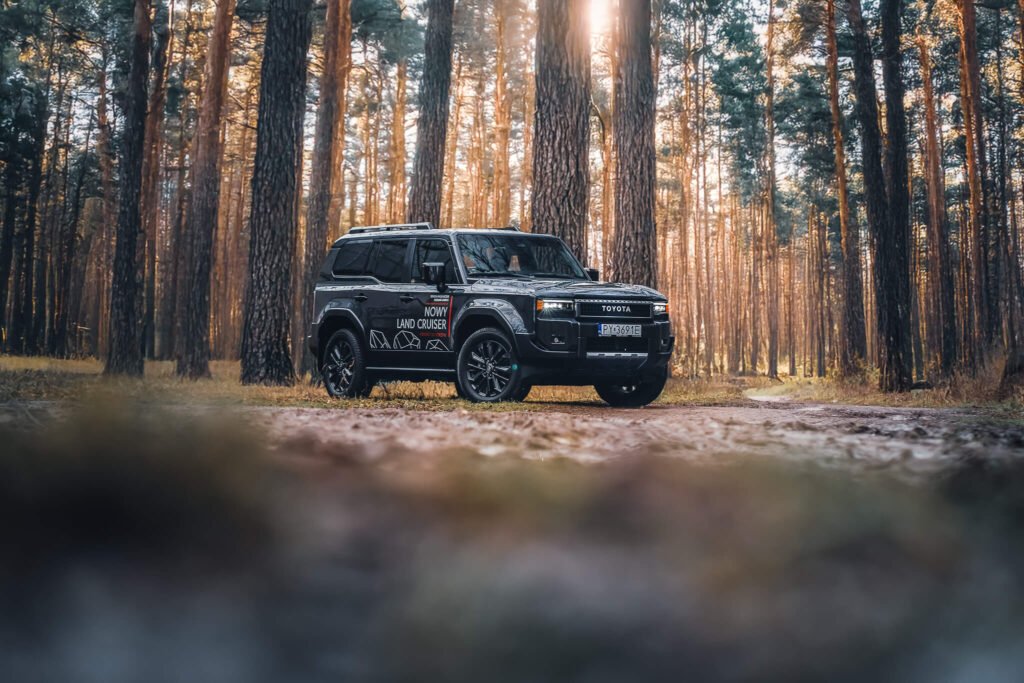 Toyota Land Cruiser w lesie, zaparkowana na leśnej drodze. Samochód wyróżnia się nowoczesnym designem, czarnym lakierem i terenowym charakterem. W tle widoczne są wysokie drzewa sosnowe, które podkreślają naturalne otoczenie i przeznaczenie pojazdu do jazdy w trudnym terenie. Idealne ujęcie dla miłośników off-roadu i przygód w plenerze.